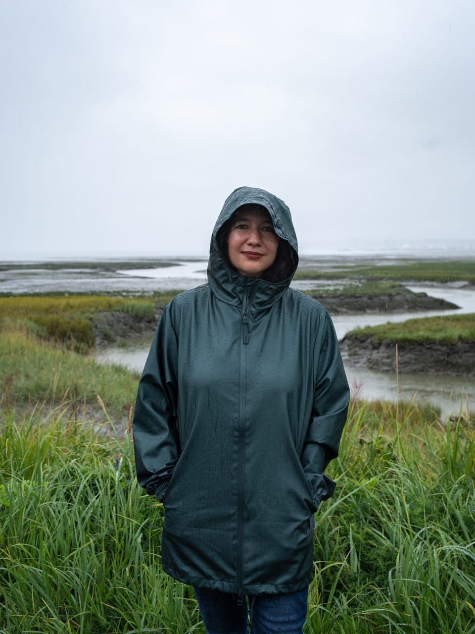 A woman stands in front of a lagoon in a raincoat while rain falls